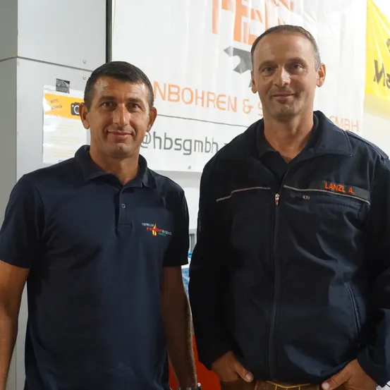 Zwei Männer stehen nebeneinander vor einem weißen Banner. Einer trägt ein dunkles Poloshirt, der andere eine dunkle Jacke. Beide lächeln.