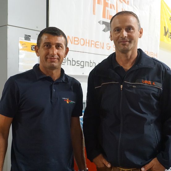 Zwei Männer stehen nebeneinander vor einem weißen Banner. Einer trägt ein dunkles Poloshirt, der andere eine dunkle Jacke. Beide lächeln.