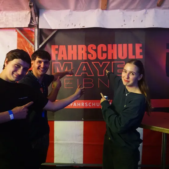 Drei Personen stehen vor einem Fahrschule Maye Elbn Banner, lächeln und posieren für die Kamera.
