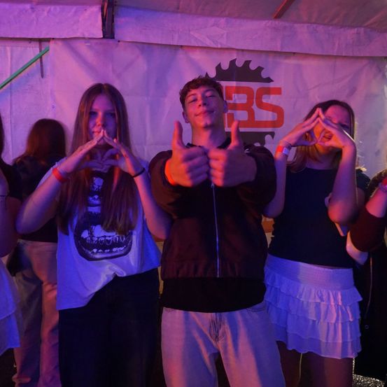 Eine Gruppe junger Leute posiert für ein Foto unter einem Zelt mit weißem Hintergrund. Ein Mädchen bildet ein Herz mit ihren Händen, ein Mann gibt einen Daumen hoch und ein anderes Mädchen bildet ein Friedenszeichen.