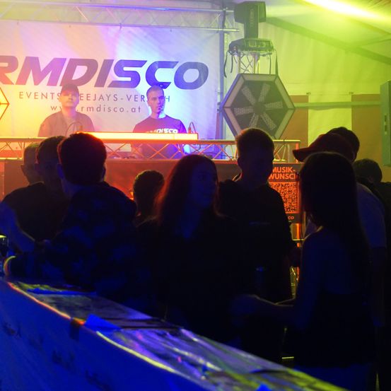 Menschen versammeln sich in einem dunklen Raum mit Neonlichtern vor einer Bühne mit zwei DJs hinter einem Tisch. Das Banner hinter der Bühne lautet 'RMDISCO'. Das Publikum scheint die Musik zu genießen.