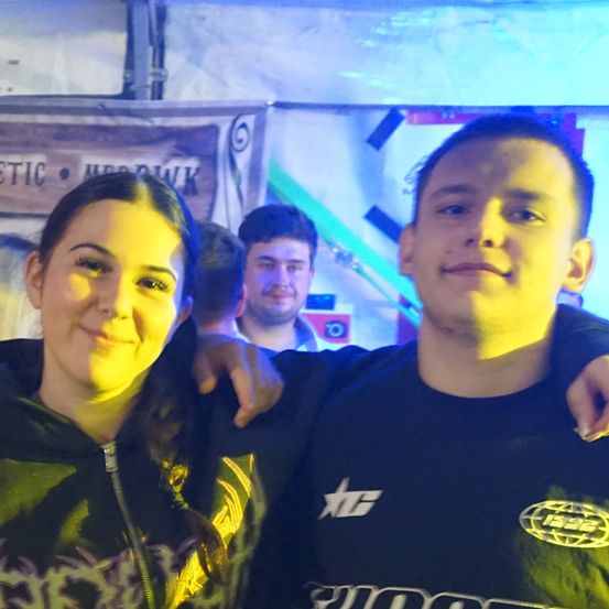 Eine lächelnde Frau und ein Mann mit dem Arm um sie, aufgenommen bei einer Veranstaltung mit bunten Lichtern und Bannern im Hintergrund.