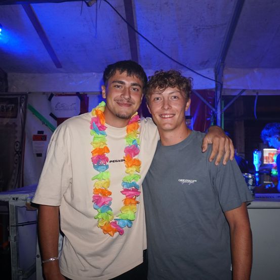 Zwei junge Männer stehen in einem Zelt, einer trägt eine bunte Lei. Der Mann auf der linken Seite hat ein Armband und ein weißes T-Shirt, während der Mann auf der rechten Seite ein graues T-Shirt trägt. Sie lächeln für die Kamera.