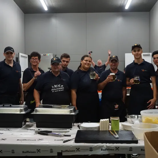 Eine Gruppe von Menschen in schwarzen Uniformen steht in einem Raum, lächelt und posiert für ein Foto. Sie halten Gläser mit Getränken. Es gibt Tabletts und Schüsseln auf dem Tisch und eine Box an der Seite. Der Raum hat weiße Wände und eine Decke mit Lichtern.