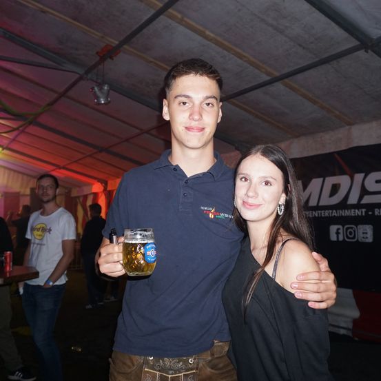 Ein junger Mann und eine junge Frau stehen lächelnd nebeneinander und halten ein Glas Bier. Sie sind lässig gekleidet, der Mann trägt ein blaues Poloshirt und die Frau ein schwarzes Top. Im Hintergrund sind mehrere Personen zu sehen, und die Umgebung wirkt nach einer lebhaften Party.
