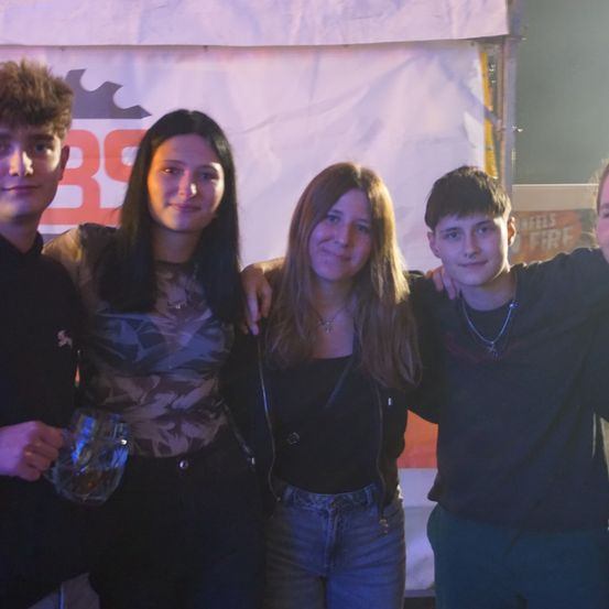 Fünf junge Leute posieren für ein Foto bei einer Veranstaltung. Die Frau auf der linken Seite hält einen Glasbecher. Ein Banner mit dem Text BS steht hinter ihnen.