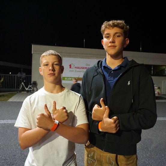 Zwei junge Männer stehen nachts vor einem Gebäude und geben beide Daumen hoch. Der Mann auf der linken Seite trägt ein weißes T-Shirt und orangefarbene Armbänder. Der Mann auf der rechten Seite trägt ein blaues Hemd und eine schwarze Jacke. Dahinter steht ein Schild mit dem Text 'Die 1. Hauskra mit Zuverl'.