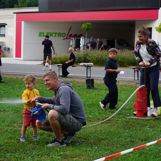 Eine Familie genießt einen Tag in Elektro Land, ein Mann sprüht Wasser aus einem Schlauch auf einen kleinen Jungen, während eine Frau hinter ihnen steht und andere Leute in der Nähe sind.