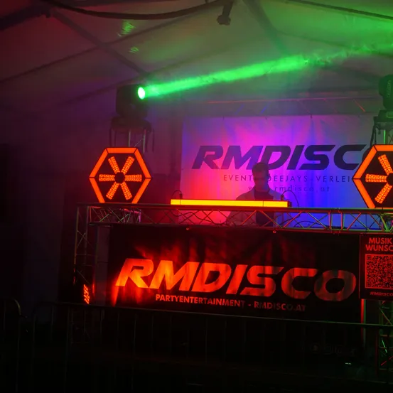 Ein DJ steht hinter einer Bühne mit RM Disco Branding, unter roten und grünen Lichtern. RM Disco ist ein Party-Entertainment-Unternehmen.
