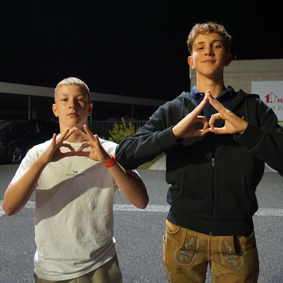 Zwei Jungen stehen nachts auf einem Parkplatz. Beide machen mit den Händen eine Herzform. Einer trägt ein weißes T-Shirt, der andere eine schwarze Kapuzenjacke. Hinter ihnen befindet sich ein Gebäude und ein geparktes Auto.