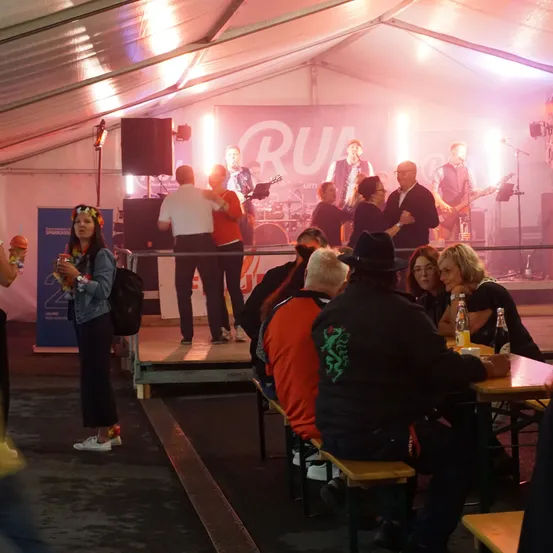Eine Band spielt auf einer Bühne in einem Zelt, vor ihnen tanzen Menschen und einige sitzen an Tischen im Vordergrund.