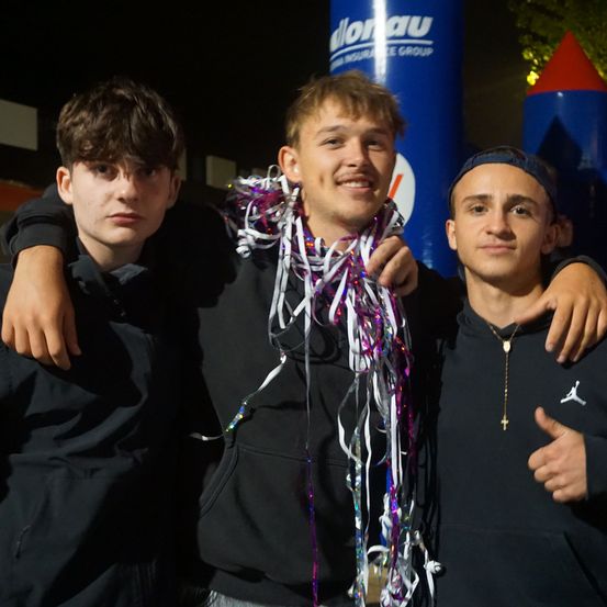 Drei junge Männer stehen eng beieinander, einer trägt eine Halskette, ein anderer hat eine Girlande um den Hals. Sie lächeln und posieren für ein Foto vor einem großen blauen Ballon.