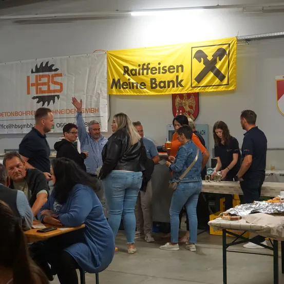Eine Gruppe von Menschen ist in einem Raum versammelt. Einige sitzen an Tischen, andere stehen. Eine Frau hält ein Glas, und ein Mann hebt die Hand. Im Hintergrund befinden sich zwei Banner, eines davon sagt Raiffeisen Meine Bank.