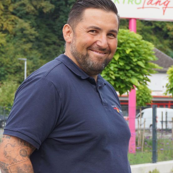 Ein Mann mit Bart und Tätowierungen am Arm steht draußen. Er trägt ein blaues Polohemd. Hinter ihm befindet sich ein rosafarbener Pfosten, Grünpflanzen und ein weißer Lieferwagen.