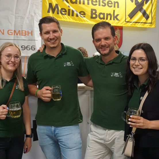 Drei Personen in grünen Poloshirts posieren mit Getränken in den Händen und lächeln vor einem Banner.