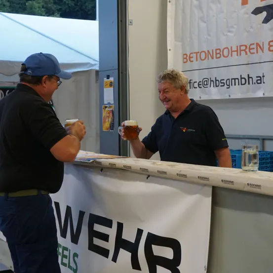Zwei Männer in Freizeitkleidung stehen hinter einem Tresen, einer lächelt und hält ein Glas Bier. Dahinter steht ein weißes Zelt und ein Banner mit orangem Text. Der Tresen hat ein weißes Banner mit schwarzem Text.