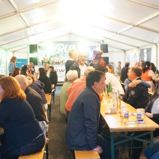 Ein Zelt beherbergt ein lebhaftes Treffen mit Menschen, die an Tischen sitzen, einige stehen und plaudern, mit einer Bühne im Hintergrund, auf der eine Band auftritt.