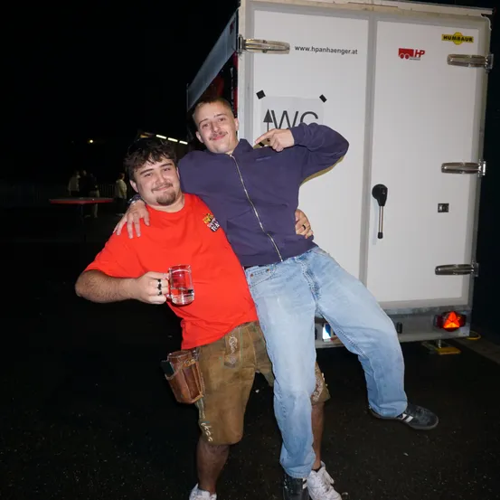 Zwei Männer stehen vor einem weißen Lastwagen bei Nacht. Ein Mann in einem roten T-Shirt hält ein Glas, der andere in einem lila Jackett.