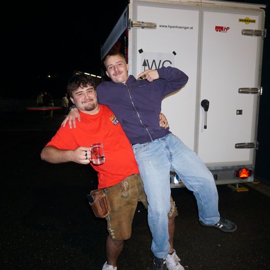 Zwei Männer stehen vor einem weißen Lastwagen bei Nacht. Ein Mann in einem roten T-Shirt hält ein Glas, der andere in einem lila Jackett.