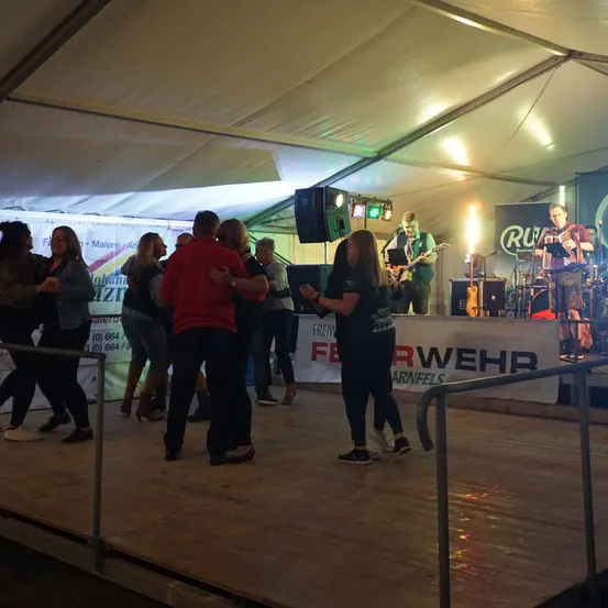 Eine Band spielt auf der Bühne in einem Zelt, während Menschen tanzen und sich unterhalten. Ein Banner mit dem Wort Wehr ist auf der Bühne sichtbar.