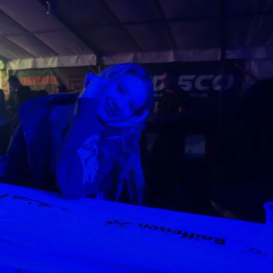 Eine Frau mit langen blonden Haaren lächelt und lehnt sich an einen blauen Tisch. Hinter ihr steht eine Person in einem dunklen Bereich mit einem Banner, das 'D 5CO' steht. Andere Menschen sind ebenfalls anwesend.