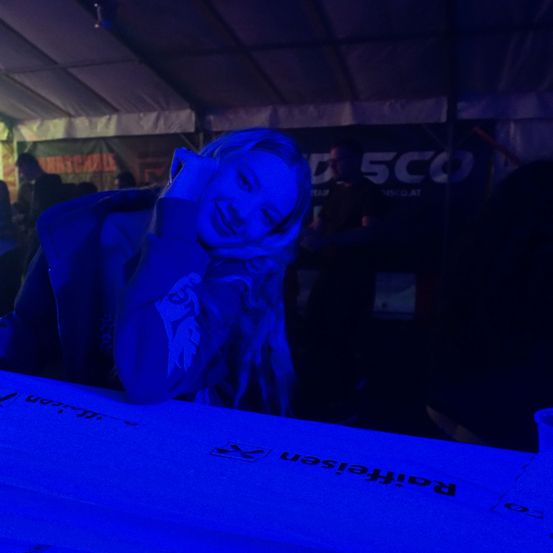 Eine Frau mit langen blonden Haaren lächelt und lehnt sich an einen blauen Tisch. Hinter ihr steht eine Person in einem dunklen Bereich mit einem Banner, das 'D 5CO' steht. Andere Menschen sind ebenfalls anwesend.