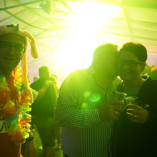 Drei Leute sind auf einer Party, einer trägt einen Hasenohren-Hut, ein anderer hält Getränke und ein Dritter küsst eine Frau. Dahinter tanzen Leute unter einem grünen Licht.