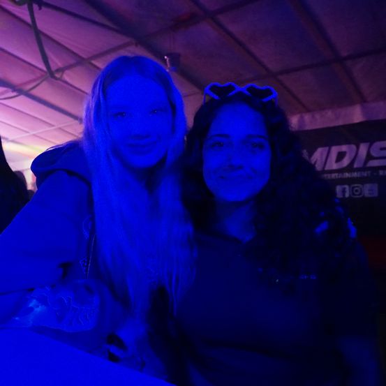 Zwei Frauen lächeln in einem dunklen Raum mit lila Beleuchtung. Eine hat lange blonde Haare und eine Kapuzenjacke, die andere hat dunkle Haare und Herzförmige Brille auf dem Kopf.