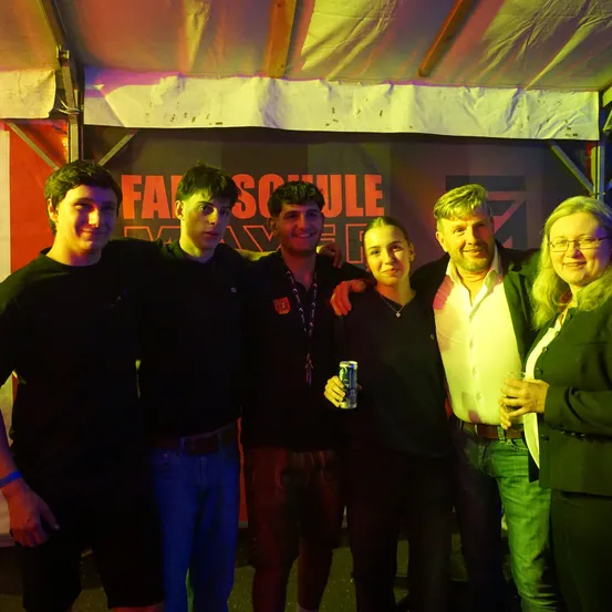 Fünf Personen posieren für ein Foto in einem Zelt mit einem Banner mit der Aufschrift 'FASSCHULE' im Hintergrund. Sie stehen unter einem mehrfarbigen Licht.