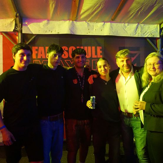 Fünf Personen posieren für ein Foto in einem Zelt mit einem Banner mit der Aufschrift 'FASSCHULE' im Hintergrund. Sie stehen unter einem mehrfarbigen Licht.