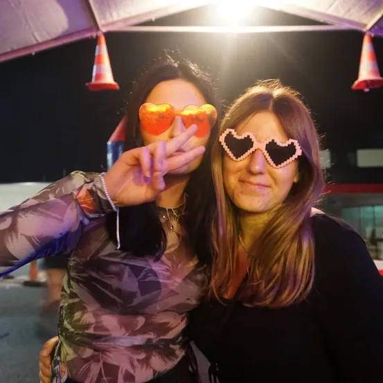 Zwei Frauen posieren nachts unter einem Zelt für ein Selfie, tragen herzförmige Sonnenbrillen und lächeln. Eine Frau hat ihre Hand in der Nähe ihres Gesichts. Hinter ihnen sind zwei orangefarbene Verkehrskegel zu sehen.