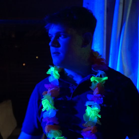 Ein Mann mit leuchtenden Blumengirlanden um den Hals steht in einem dunklen Raum mit blauer Beleuchtung.