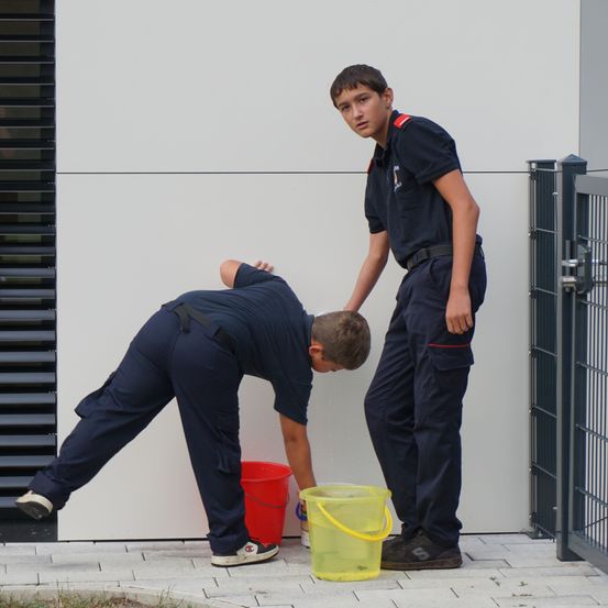 Zwei junge Männer in dunkelblauen Uniformen reinigen den Bereich vor einem Gebäude. Ein Mann ist gebeugt und scheint den Boden zu waschen, während der andere Mann hinter ihm steht.