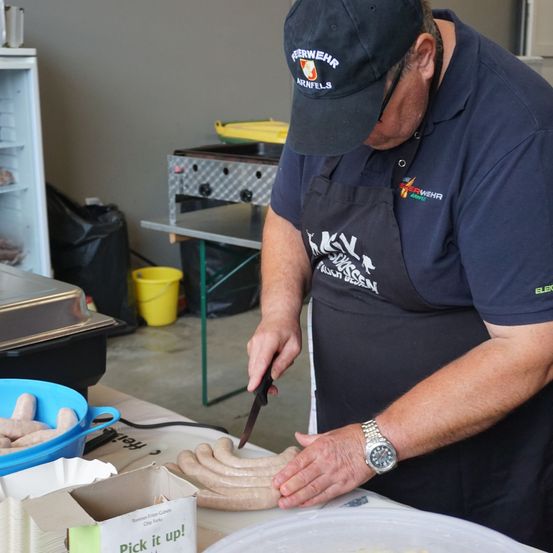 Ein Mann in einem blauen Hemd und einer Kappe schneidet Würste auf einem Schneidebrett. Er trägt eine Uhr und eine schwarze Schürze. Es gibt Würste in einer Schüssel und eine Schachtel mit dem Text Pack es ein! auf dem Tisch.