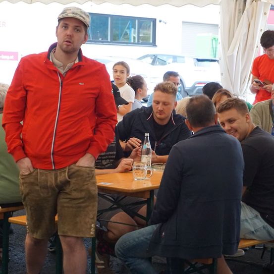 Eine Gruppe von Menschen sitzt um einen Tisch in einem Zelt im Freien, ein Mann steht in der Nähe von ihnen und ein Mädchen schaut auf ein Telefon.