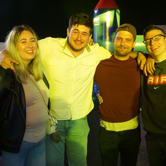 Vier Personen lächeln und posieren für ein Foto bei Nacht. Ein Mann in einem gelben Hemd und grüner Hose steht zwischen zwei anderen Männern. Der Mann auf der rechten Seite trägt eine Brille und ein Nike-Shirt. Hinter ihnen steht ein großer aufblasbarer Gegenstand.