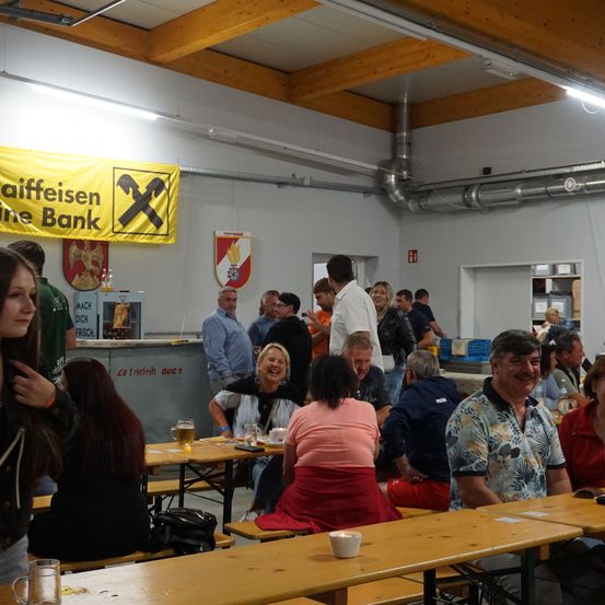 Eine Gruppe von Menschen sitzt und steht um Tische in einem Raum mit Holzbalken, einige trinken aus Gläsern. Banner hängen an der Wand.