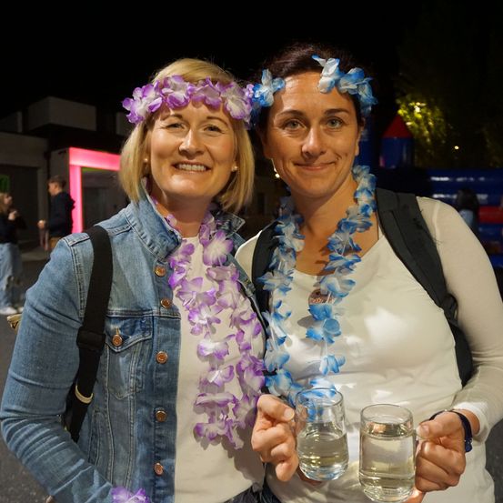 Zwei Frauen mit Blumenkränzen halten Gläser mit Wasser in der Hand und lächeln. Eine Frau trägt eine Jeansjacke. Im Hintergrund ist ein Gebäude mit einem rosa Eingang und Leute zu sehen.