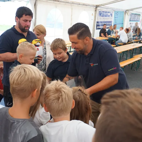 Eine Gruppe von Kindern ist um einen Mann in einem blauen Hemd versammelt, der ihnen etwas in einem Glaskasten zeigt. Dahinter stehen eine Frau und ein Mann.