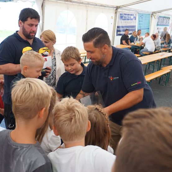 Eine Gruppe von Kindern ist um einen Mann in einem blauen Hemd versammelt, der ihnen etwas in einem Glaskasten zeigt. Dahinter stehen eine Frau und ein Mann.