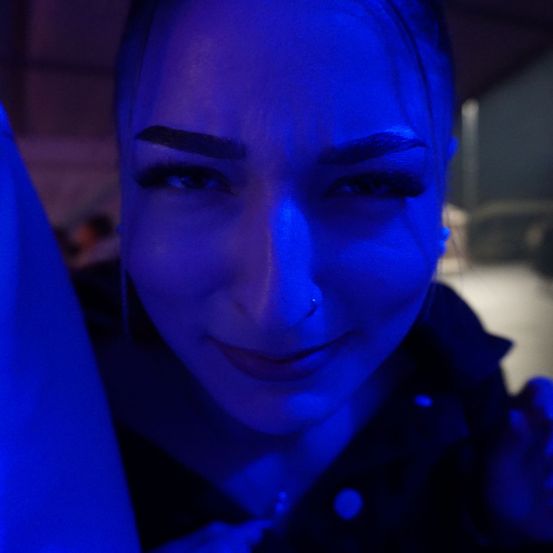Eine Frau lächelt mit blauem Licht, das ihr Gesicht beleuchtet. Sie hat hochgezogene Augenbrauen, einen Nasenring und einen leicht geöffneten Mund.