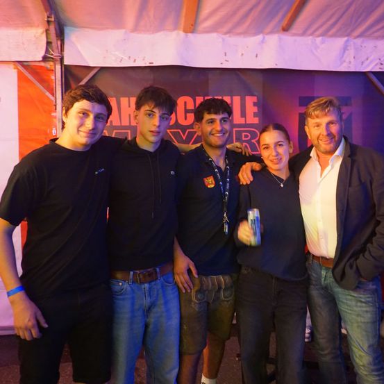 Fünf Personen posieren vor einem Schild, das 'AHCLE' steht. Die beiden rechts tragen formale Kleidung. Der Mann links hält eine Dose. Die beiden in der Mitte tragen Freizeitkleidung.