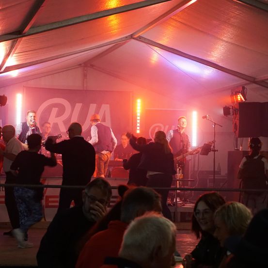 Eine Band spielt auf der Bühne unter einem Zelt mit roter und lila Beleuchtung. Das Publikum, einige stehend und andere sitzend, beobachtet mit Begeisterung. Die Bühne ist mit einem Banner mit dem Wort 'RUA' und zusätzlichen Beleuchtungskörpern geschmückt.
