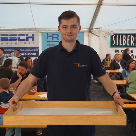 Ein Mann in einem schwarzen Shirt hält ein hölzerne Tablett in einem überfüllten Außenbereich. Im Hintergrund sitzen mehrere Personen an Tischen unter einem Zelt. Banner mit den Texten 'ESCH Transporte' und 'Silber' sind sichtbar.