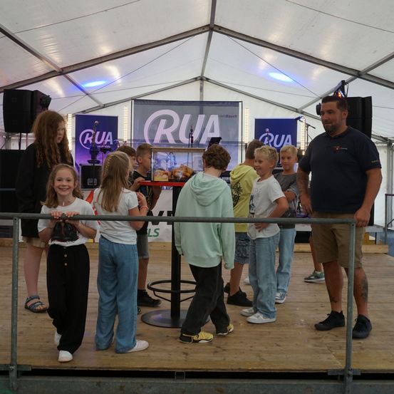 Eine Gruppe von Kindern und ein Mann stehen auf einer Bühne, wahrscheinlich bei einer Veranstaltung. Sie sind in der Nähe eines Tisches mit Snacks und einem Mikrofon. Die Bühne hat ein weißes Zeltdach und Banner mit den Buchstaben RUA.