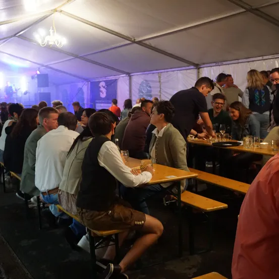 Eine große Gruppe von Menschen sitzt an Tischen in einem Zelt. Die Tische haben Getränke und Essen darauf. Einige Leute stehen. Ein bärtiger Mann steht hinter dem Tisch und spricht mit einer Frau. Ein Mann in einer Weste sitzt auf einer Bank und schaut die Frau an.
