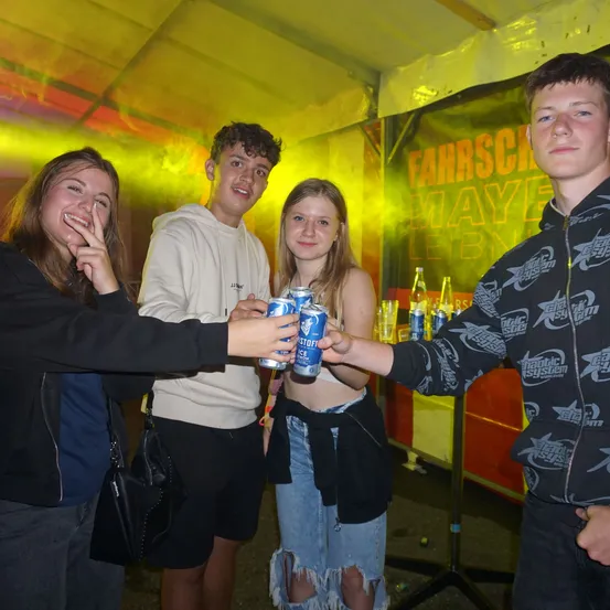 Vier junge Leute stehen lächelnd in einem Zelt. Zwei halten Dosen Eiskaltes Bier. Ein Banner mit der Aufschrift 'Fahrschule Maye Leben'.