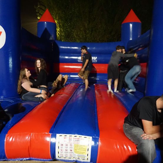 Mehrere Personen befinden sich in einem blau-roten aufblasbaren Hüpfburg. Einige sitzen, andere stehen, eine Person kniet auf dem Boden. Eine Sicherheitstafel ist an der Seite angebracht.