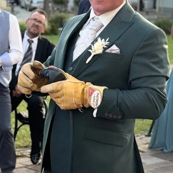 Ein Mann in einem grünen Anzug hält eine Flasche in gelben Handschuhen. Er trägt eine weiße Blume und eine Krawatte. Hinter ihm sitzen zwei Männer auf einer Bank.