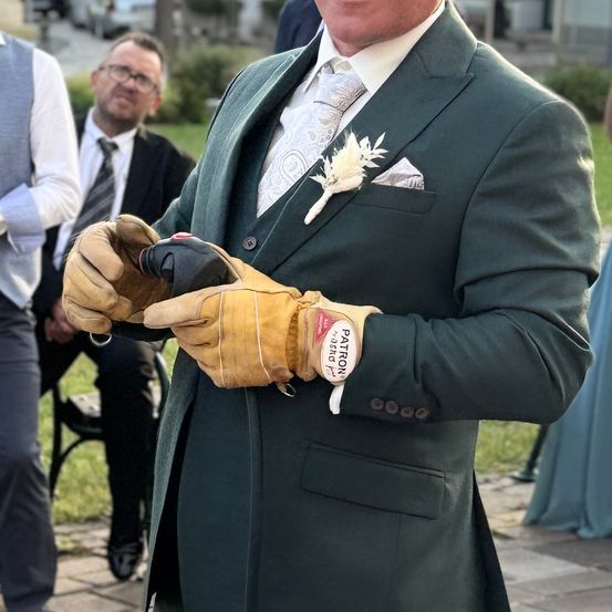 Ein Mann in einem grünen Anzug hält eine Flasche in gelben Handschuhen. Er trägt eine weiße Blume und eine Krawatte. Hinter ihm sitzen zwei Männer auf einer Bank.